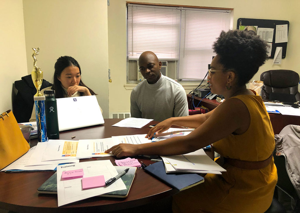 Three people sit around a table covered in documents in an office. One person points to a paper while talking to the others.