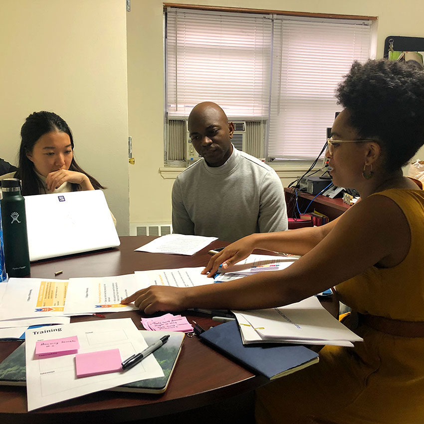 Three people sit around a table covered in documents in an office. One person points to a paper while talking to the others.