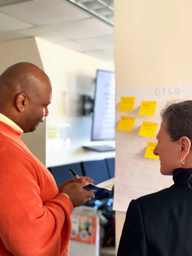 Two individuals facing a wall of yellow post-its notes while one individual takes notes in a notebook.