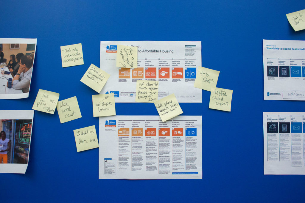 A printed out graphic of what to expect while seeking affordable housing is taped to a blue wall. Several sticky notes cover the wall and printout with notes about how to improve the graphic.