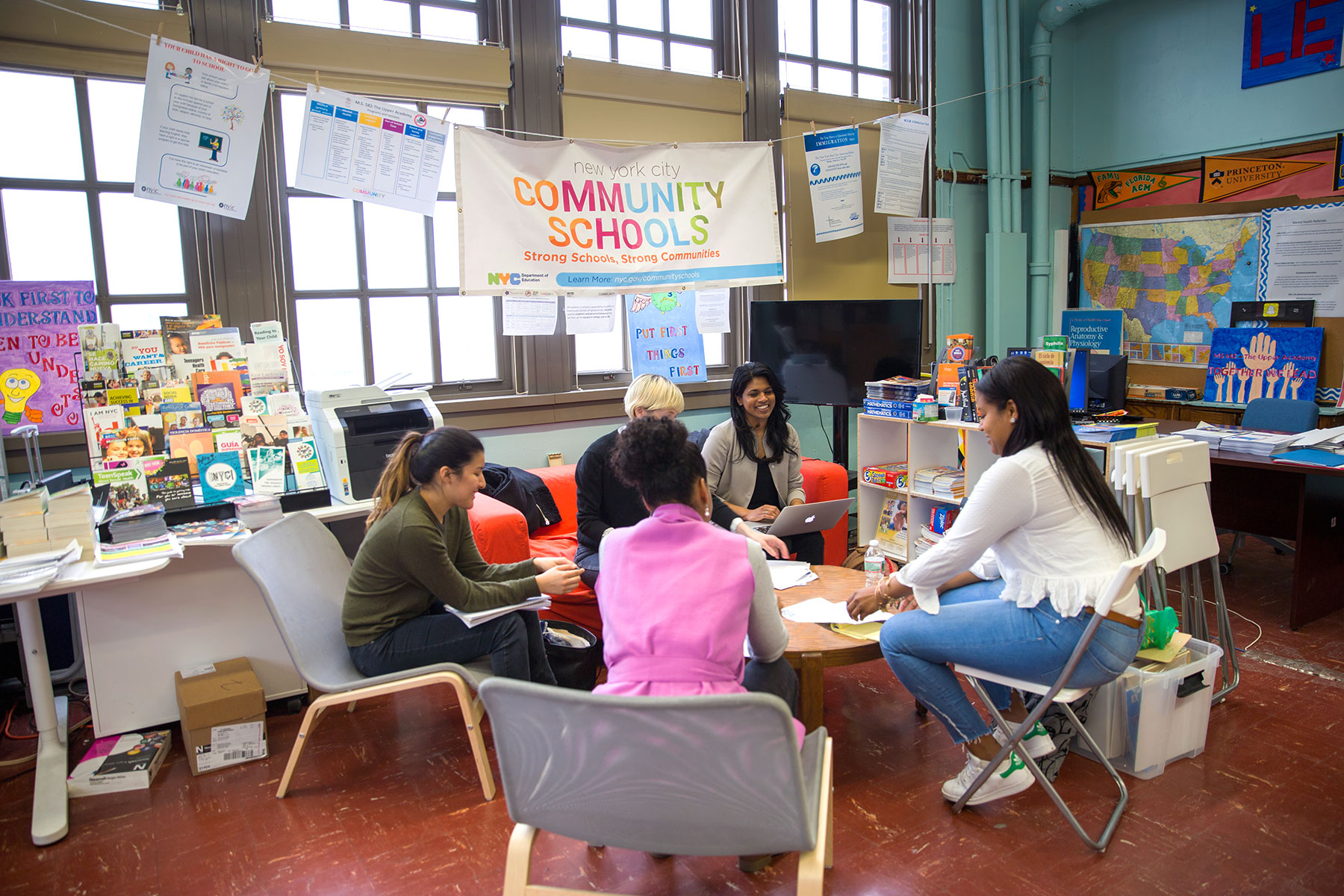 Public Policy Lab team members talk around a table with collaborators in a NYC public school. A community schools banner hangs behind them in a room full of books and children's art.