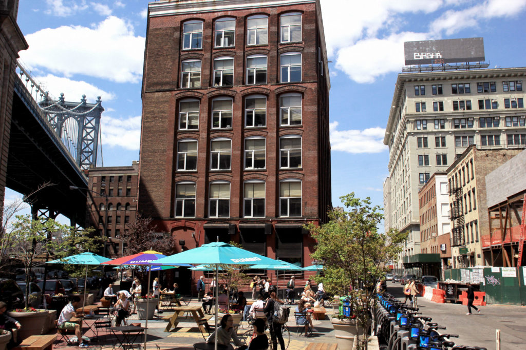 People sit at picnic tables in Dumbo Square with the Brooklyn Bridge, brick buildings, and Citi Bikes surrounding them.