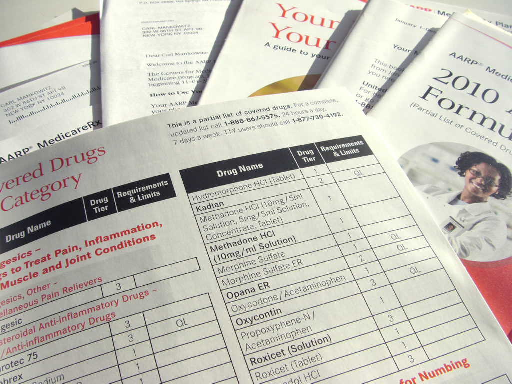 A stack of old Medicare guides with one on top open to a page of drugs covered by insurance.