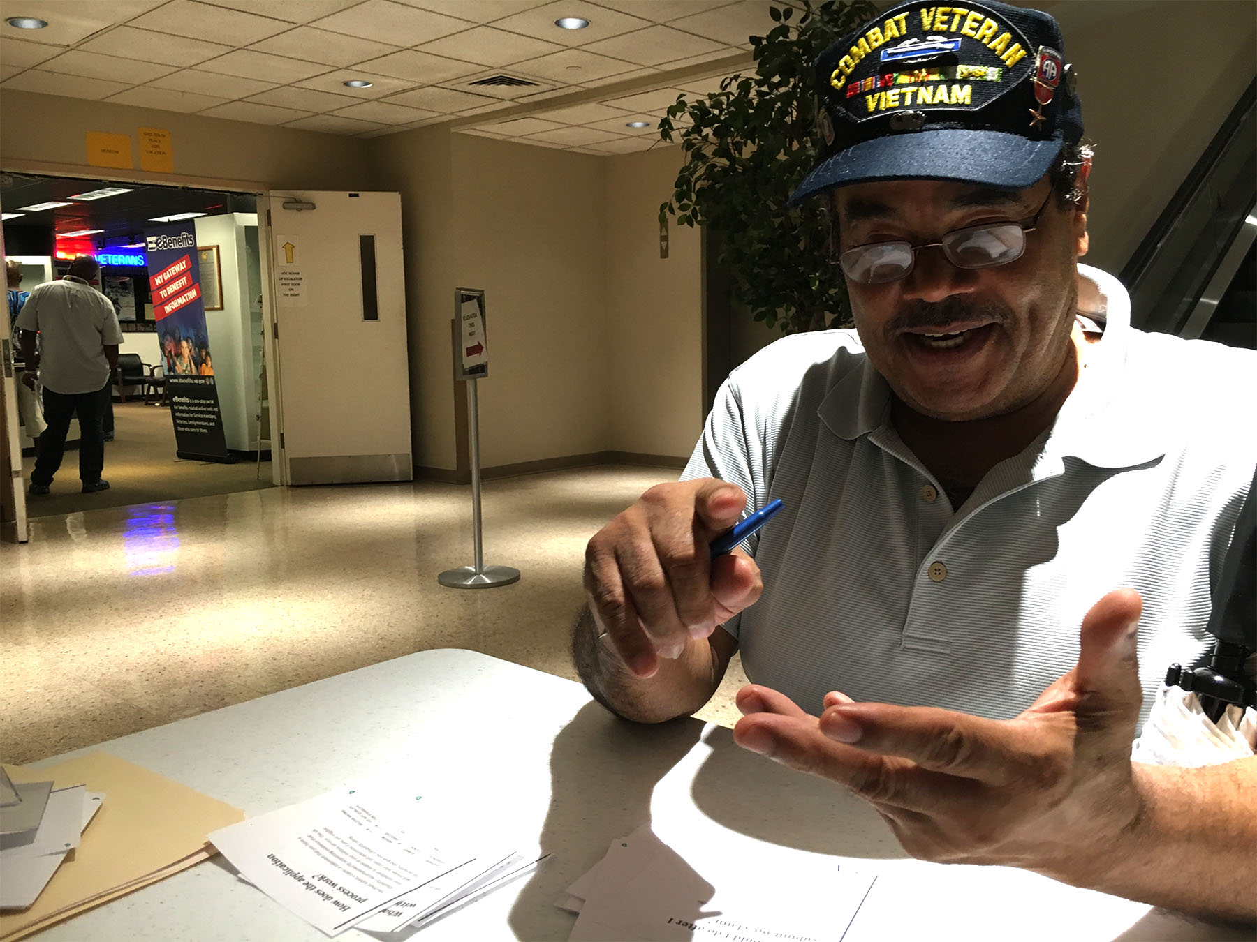 A person in a "Combat Veteran: Vietnam" hat gestures with his hands while talking at a table.