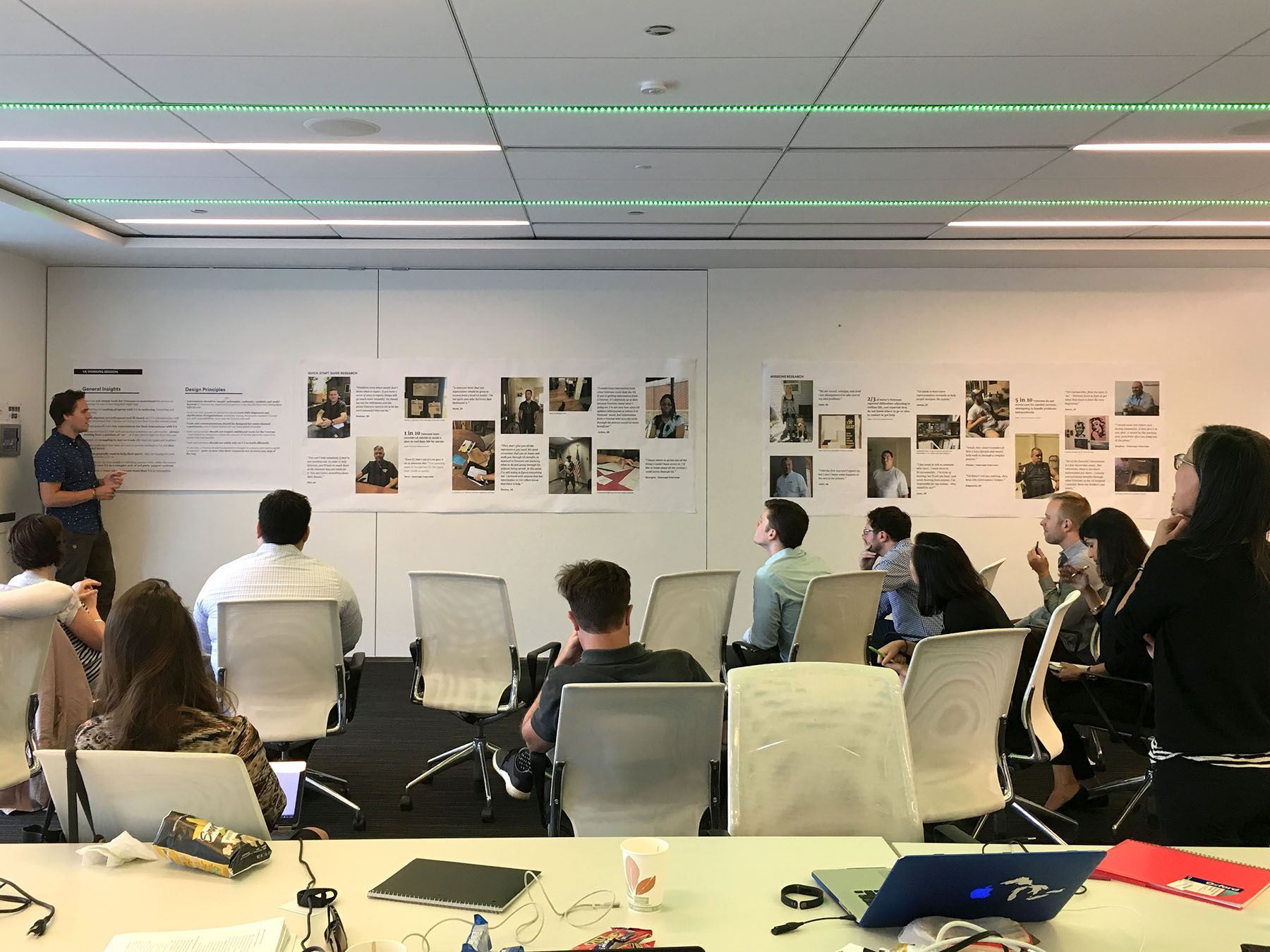A group of people sit in an office watching a presenter discuss several large posters of research materials on the wall.