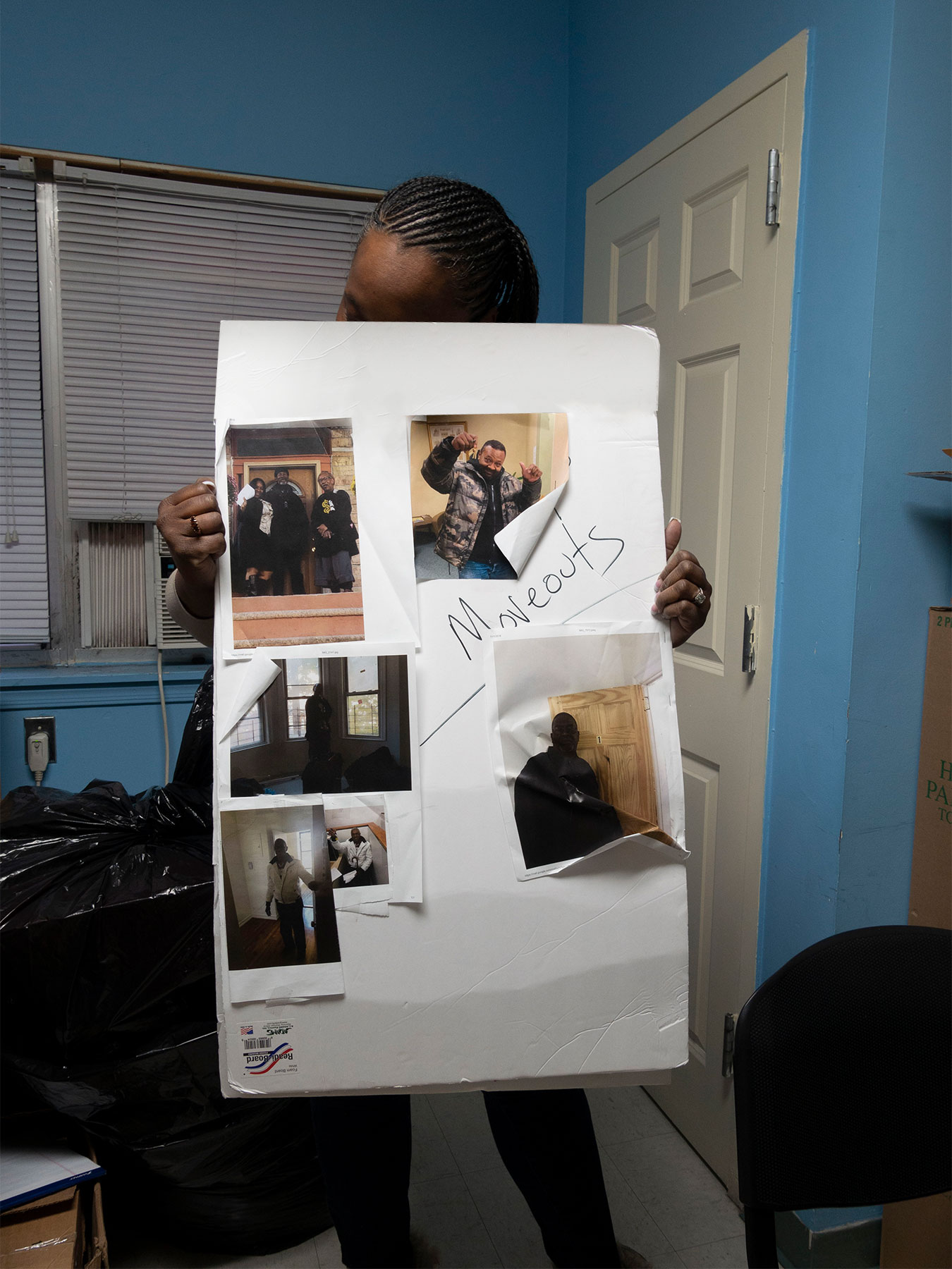 An individual holds up a poster paper covered in photographs of other people with the words "move outs" written in large in black marker.