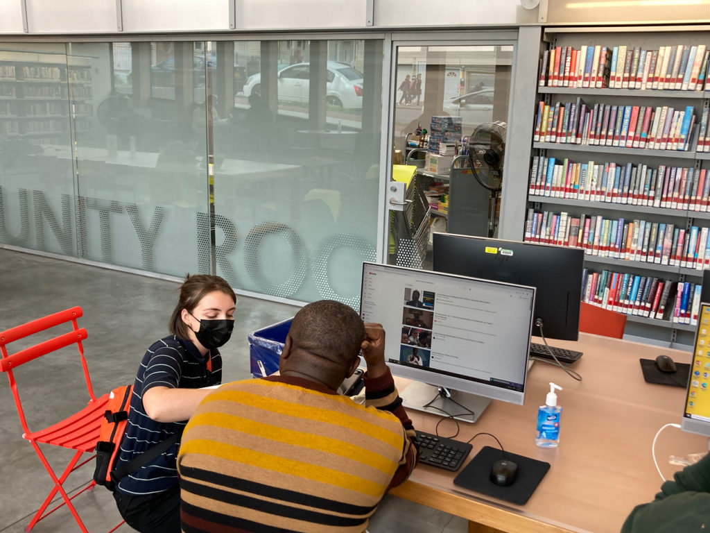 A PPL team member kneels next to a participant seated at a library computer and writes notes.