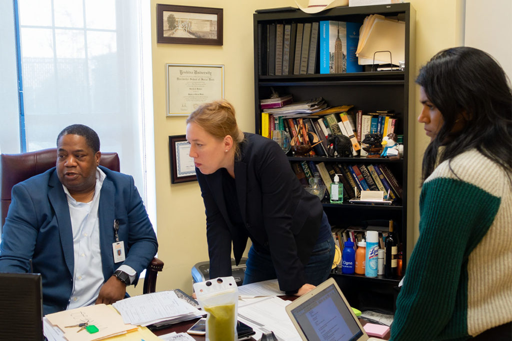 Two PPL team members stand at the desk of a collaborator while he shows them something on his computer screen.