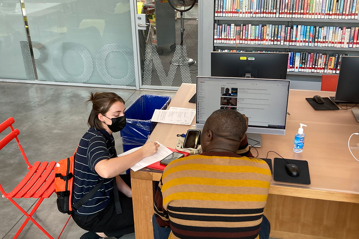 A PPL team member fills out a form while kneeling next to a collaborator who is seated at a library computer.