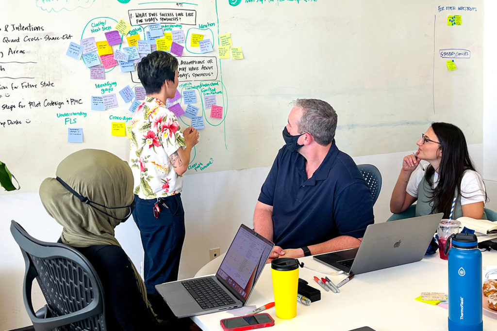 Four people face a wall covered in sticky notes during a research synthesis session. One stands at the wall reading from the stickies while the others sit in front of her.