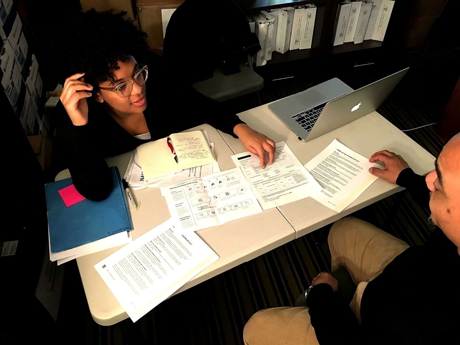 Two individuals sit across from each other at a table. They are in conversation while looking at several papers laid out in front of them.