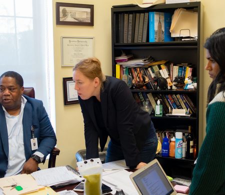 Two PPL team members stand at the desk of a collaborator while he shows them something on his computer screen.
