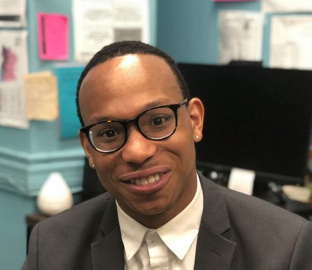 A person in a suit smiles, facing the a camera in front of a computer and a wall covered in taped-up documents.